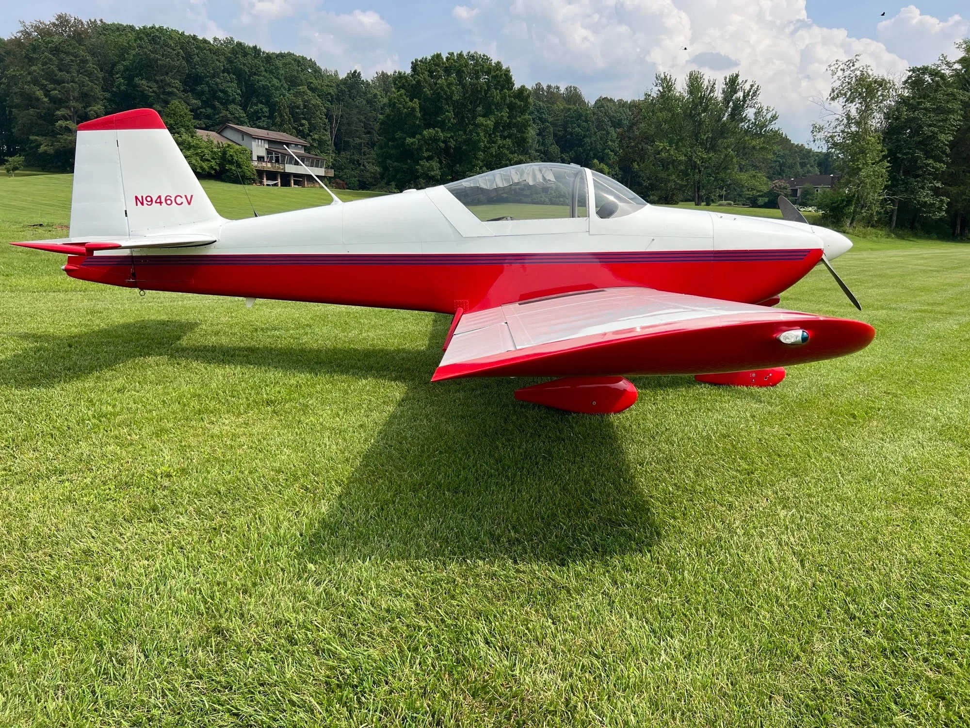 A white and red single-engine aircraft with tail number N946CV is parked on a grassy field under a partly cloudy sky.