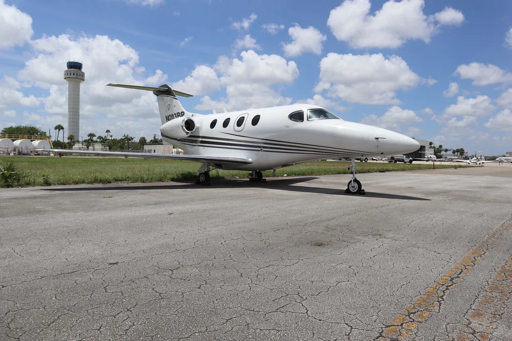 A white Embraer Phenom 100 business jet with black and gold stripes, parked on an asphalt ramp with an airport control tower in the background.