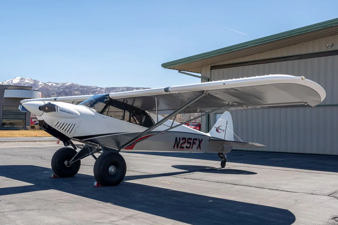 A three-quarter front view of the white and grey Carbon Cub FX-3 aircraft N25FX parked on an asphalt ramp next to a hangar.