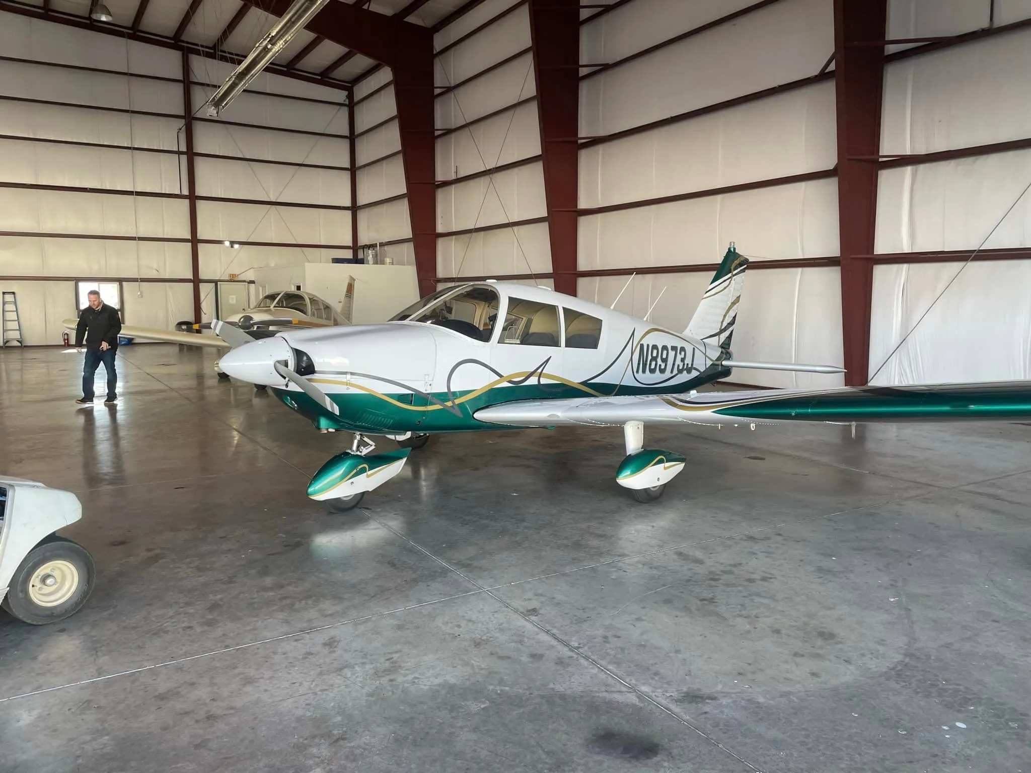 A white and green single-engine aircraft with gold accents, tail number N8973J, parked inside a hangar.