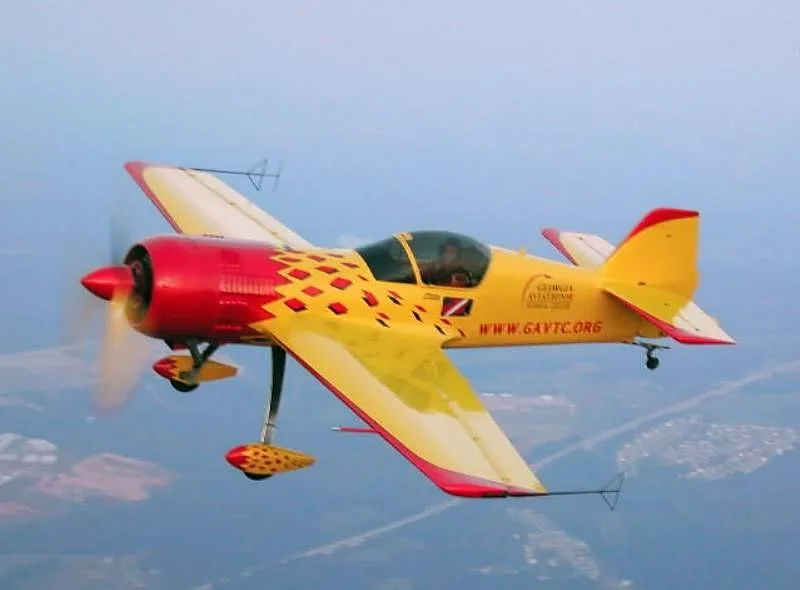 A yellow and red Sukhoi Su-26 aerobatic aircraft with 'GEORGIA AVIATION TECHNICAL COLLEGE' markings, flying against a hazy blue sky.