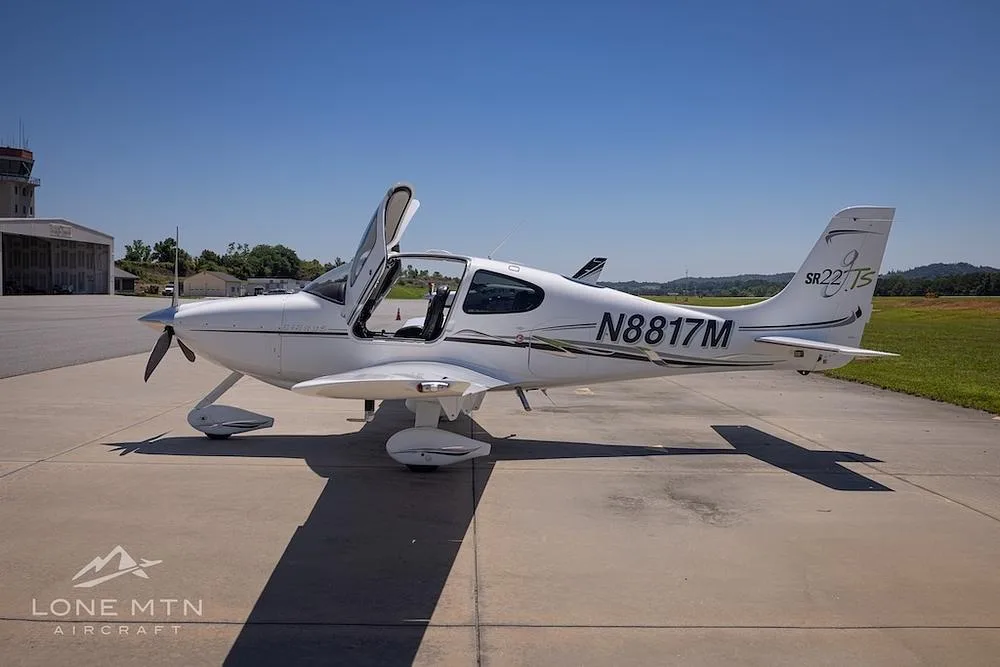 A white Cirrus SR22T aircraft (N8817M) with doors open, parked on a tarmac with a control tower in the background.