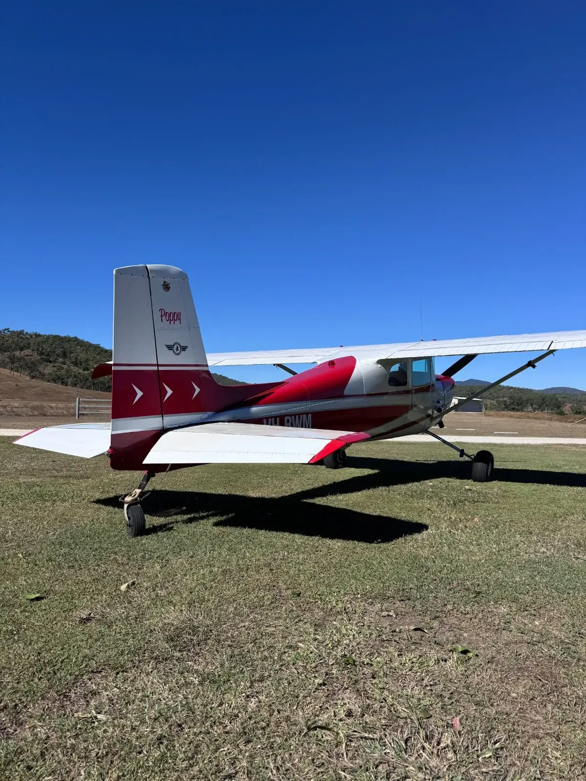 A rear-side view of a red and white single-engine propeller aircraft, registration VH-RWM, on a grassy field.