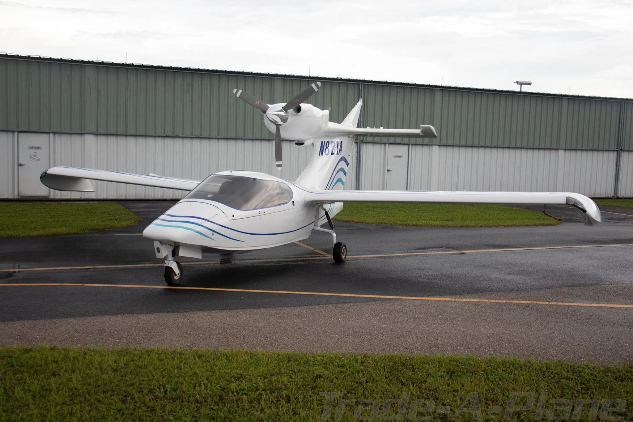 A front-left view of a white Searey amphibian aircraft with blue stripes, registration N821A, parked on a wet tarmac.