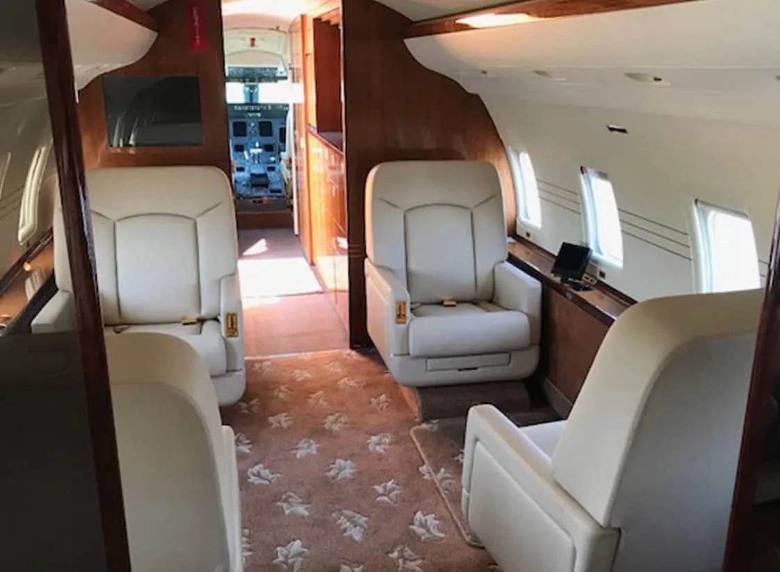 The interior of a private jet cabin features cream leather seats, wood paneling, and a patterned carpet, with a view into the cockpit.