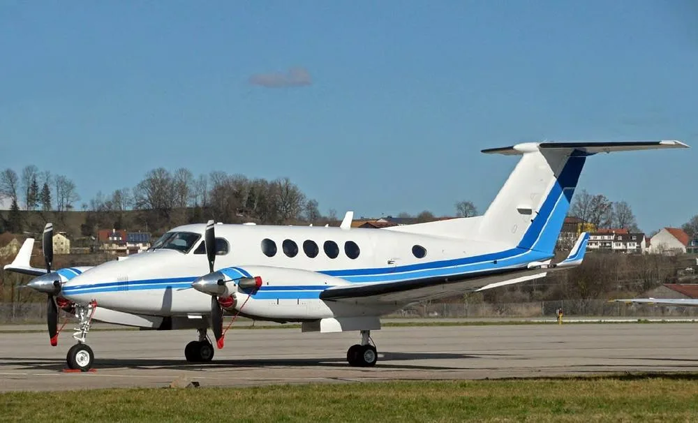A white twin-turboprop aircraft with blue stripes on the fuselage and tail, parked on an airfield under a clear sky.