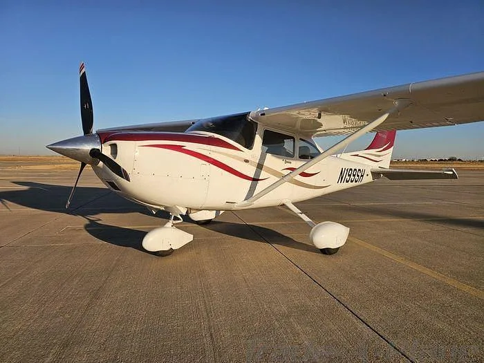 A white Cessna 182 with red and maroon stripes is parked on an asphalt runway under a clear blue sky.