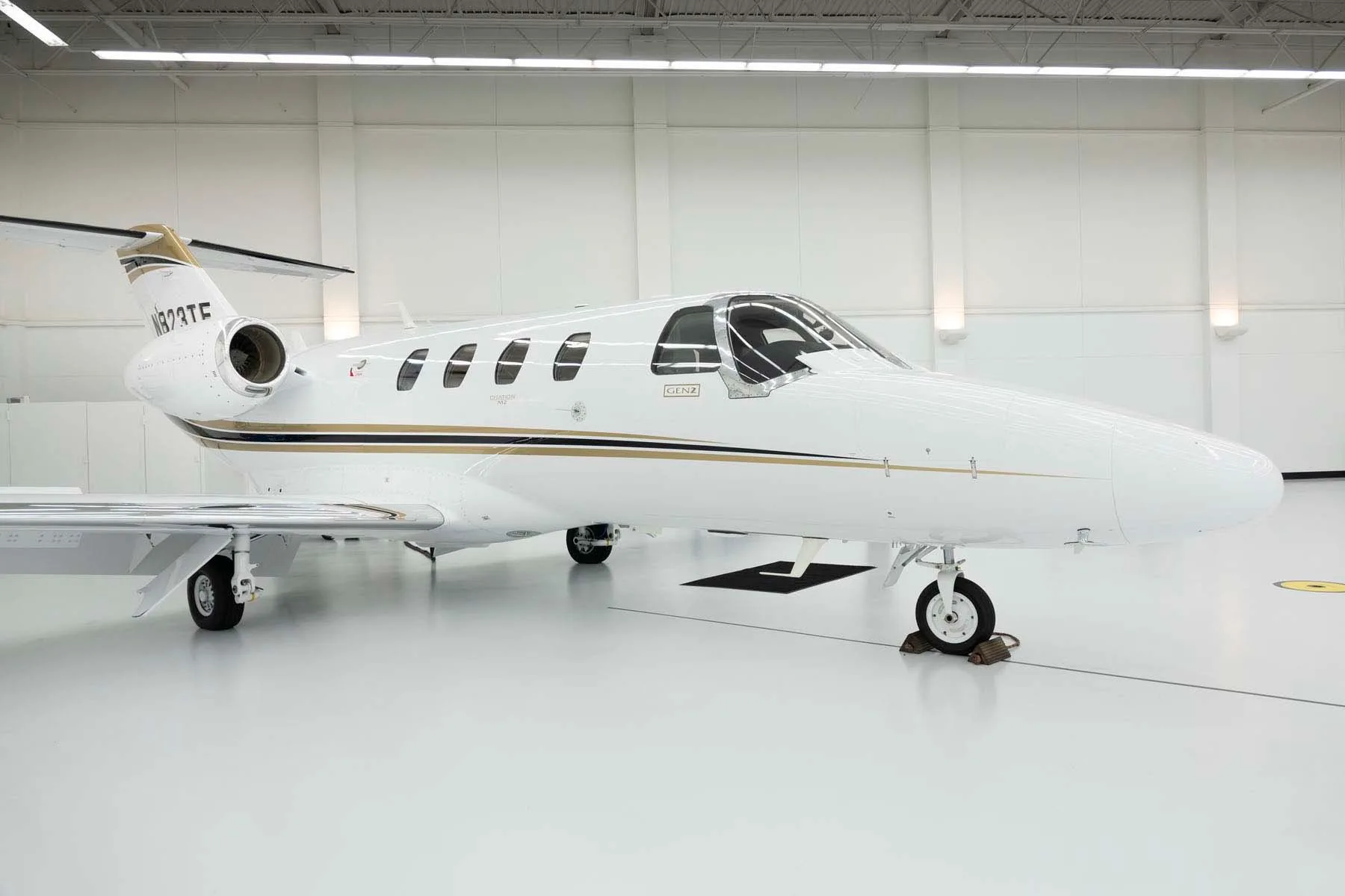 A white Cessna Citation M2 jet with gold and black stripes, parked in a brightly lit hangar.
