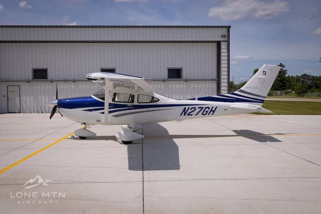 A side view of a white and blue Cessna 182 Skylane (N27GH) parked on an airport tarmac next to a hangar.