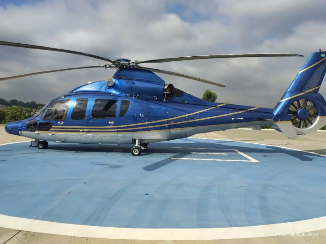 A blue and silver helicopter with gold stripes is parked on a blue helipad, viewed from the left side.