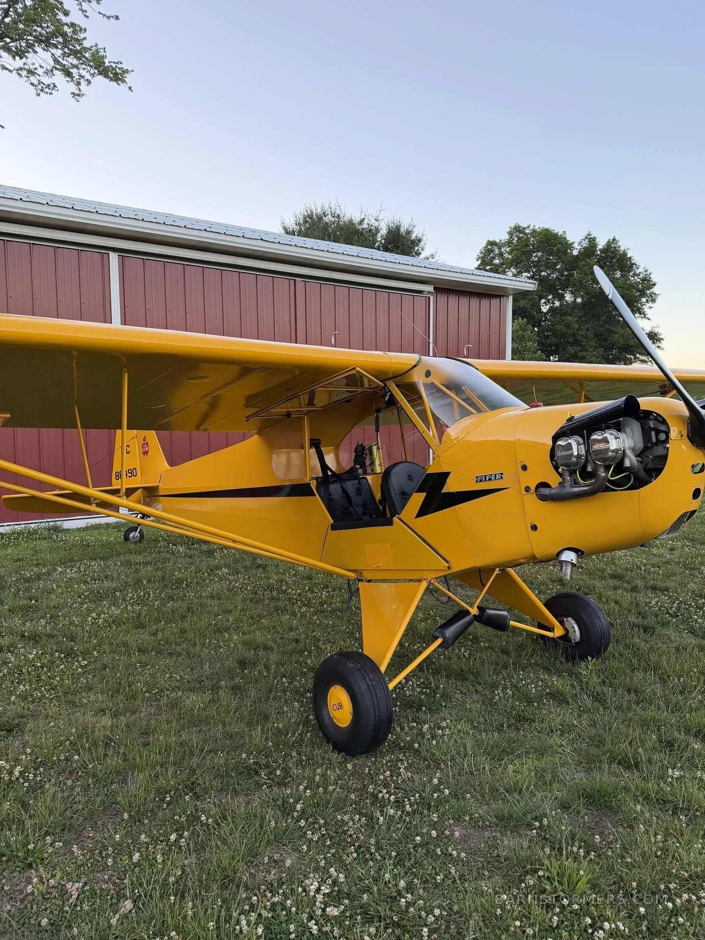 A yellow Piper Cub aircraft with its engine exposed is parked on a grassy field next to a red building.