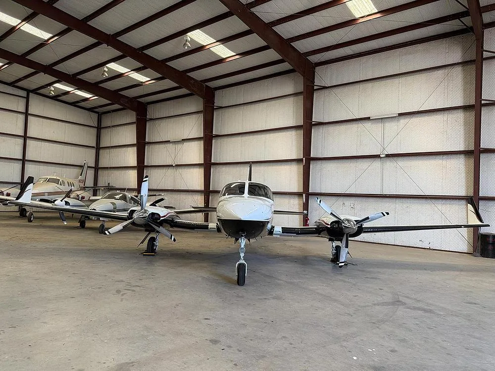 The twin-engine aircraft N414SM, with white and black paint and gold trim, parked head-on in a hangar with other planes.