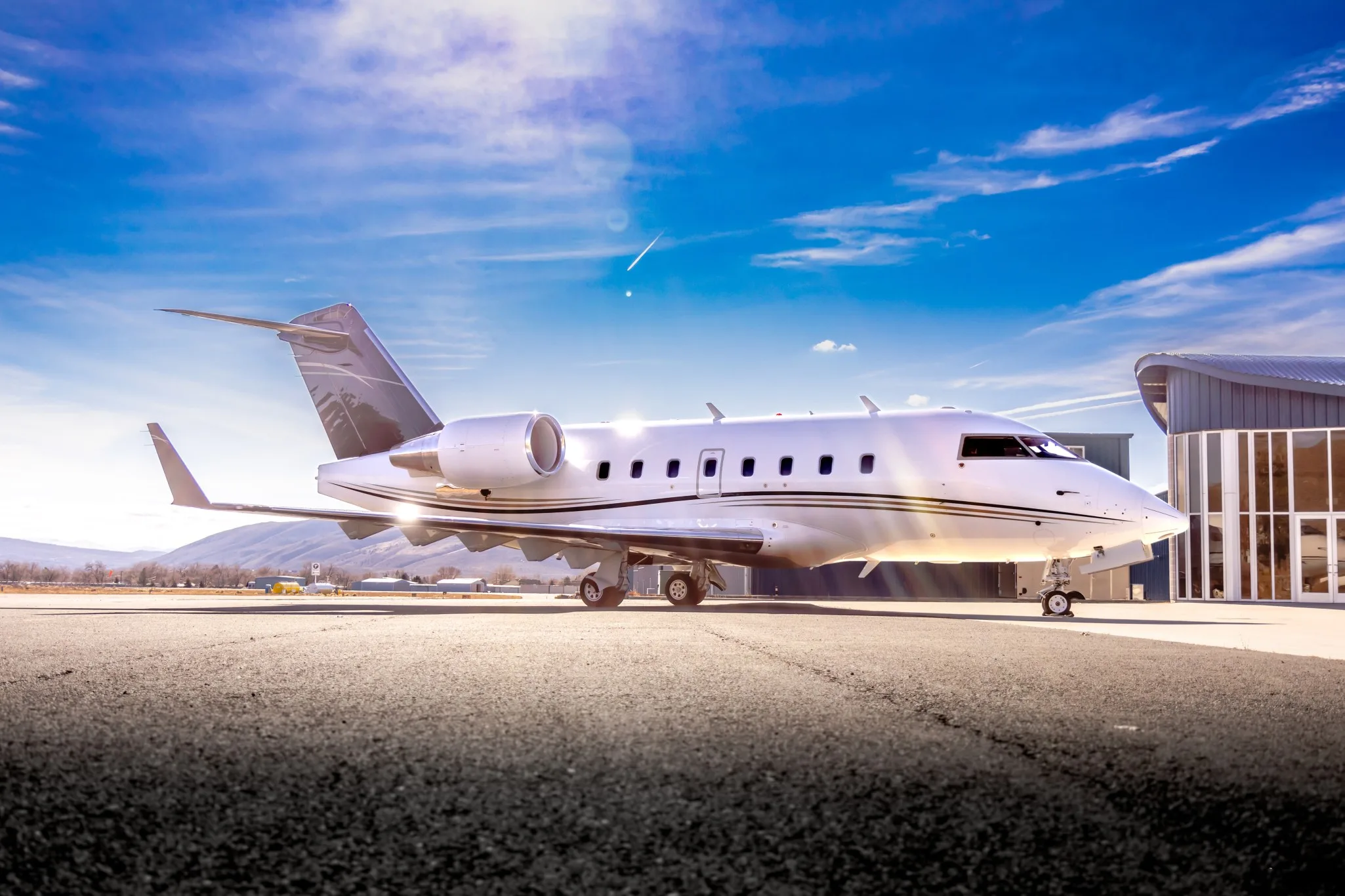 A white Bombardier Challenger business jet is parked on a tarmac with a modern hangar and mountains under a bright sky.