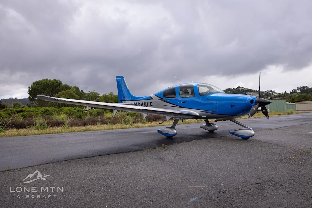 A side view of a blue and silver single-engine propeller aircraft (Cirrus SR22) with registration N248LF parked on a wet tarmac under a cloudy sky.