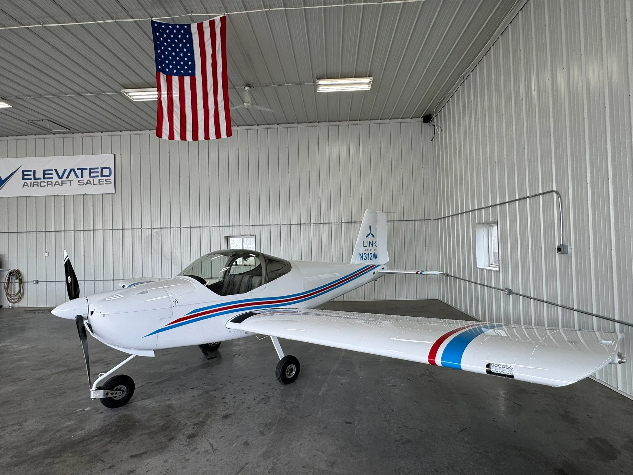 A white Van's RV-12iS aircraft with red and blue stripes on its fuselage and wings, parked in a hangar with an American flag hanging.