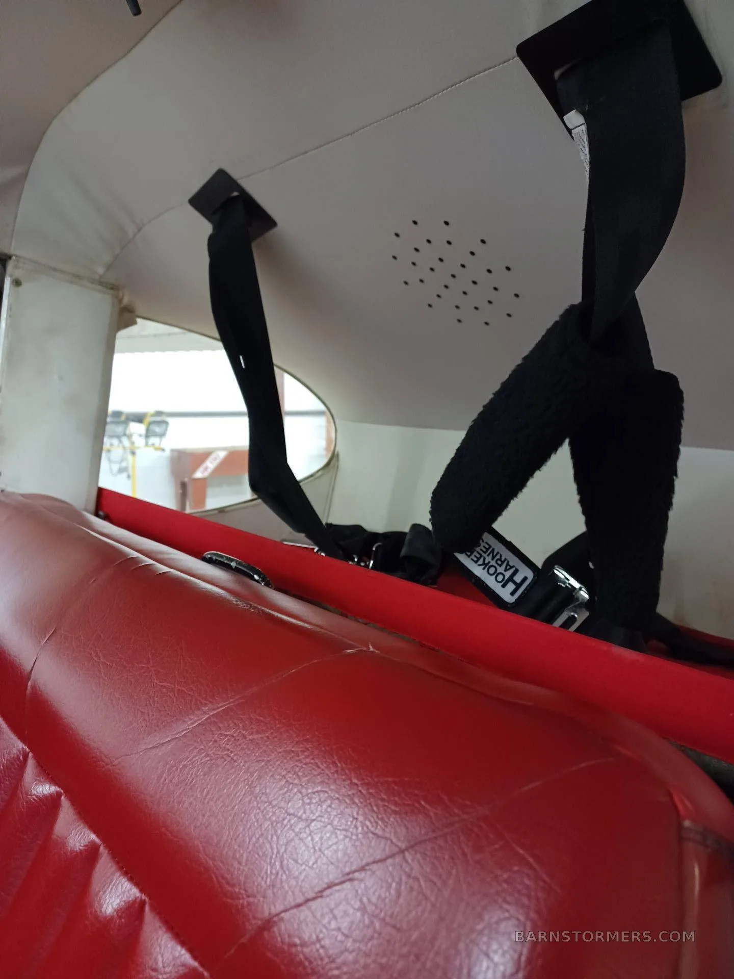 The interior cabin of a small aircraft showing red upholstered seats and black shoulder harnesses attached to the ceiling.