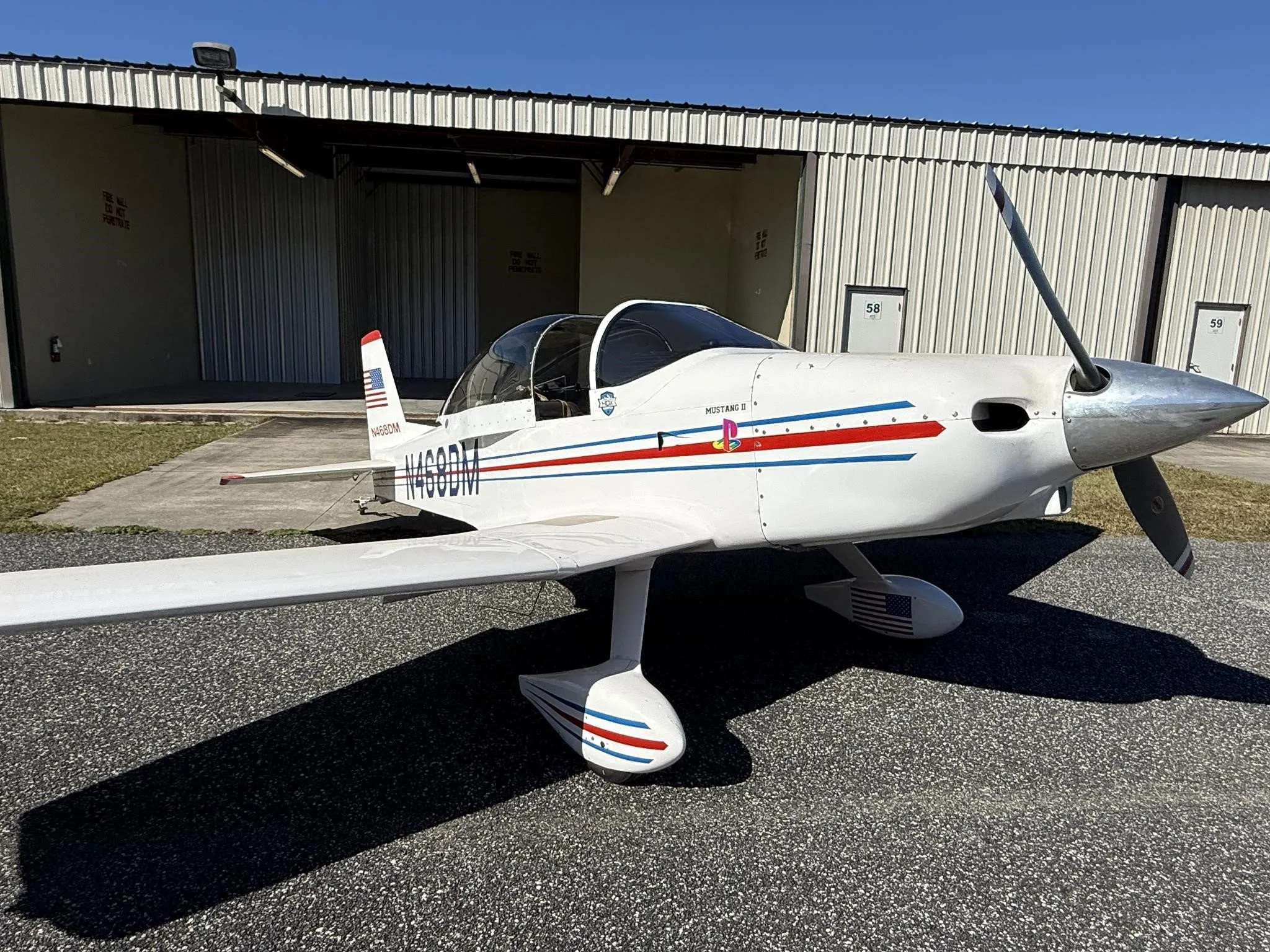 A white Mustang II aircraft with red and blue stripes, registration N468DM, parked on a tarmac in front of hangars.