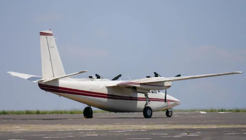 A white twin-engine aircraft with a maroon stripe on a tarmac, viewed from the rear-left.
