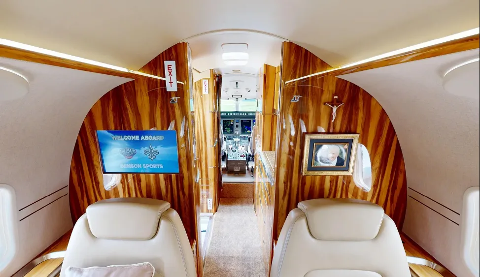 The luxurious interior of a private jet cabin with light leather seats, wood paneling, and a view into the cockpit.