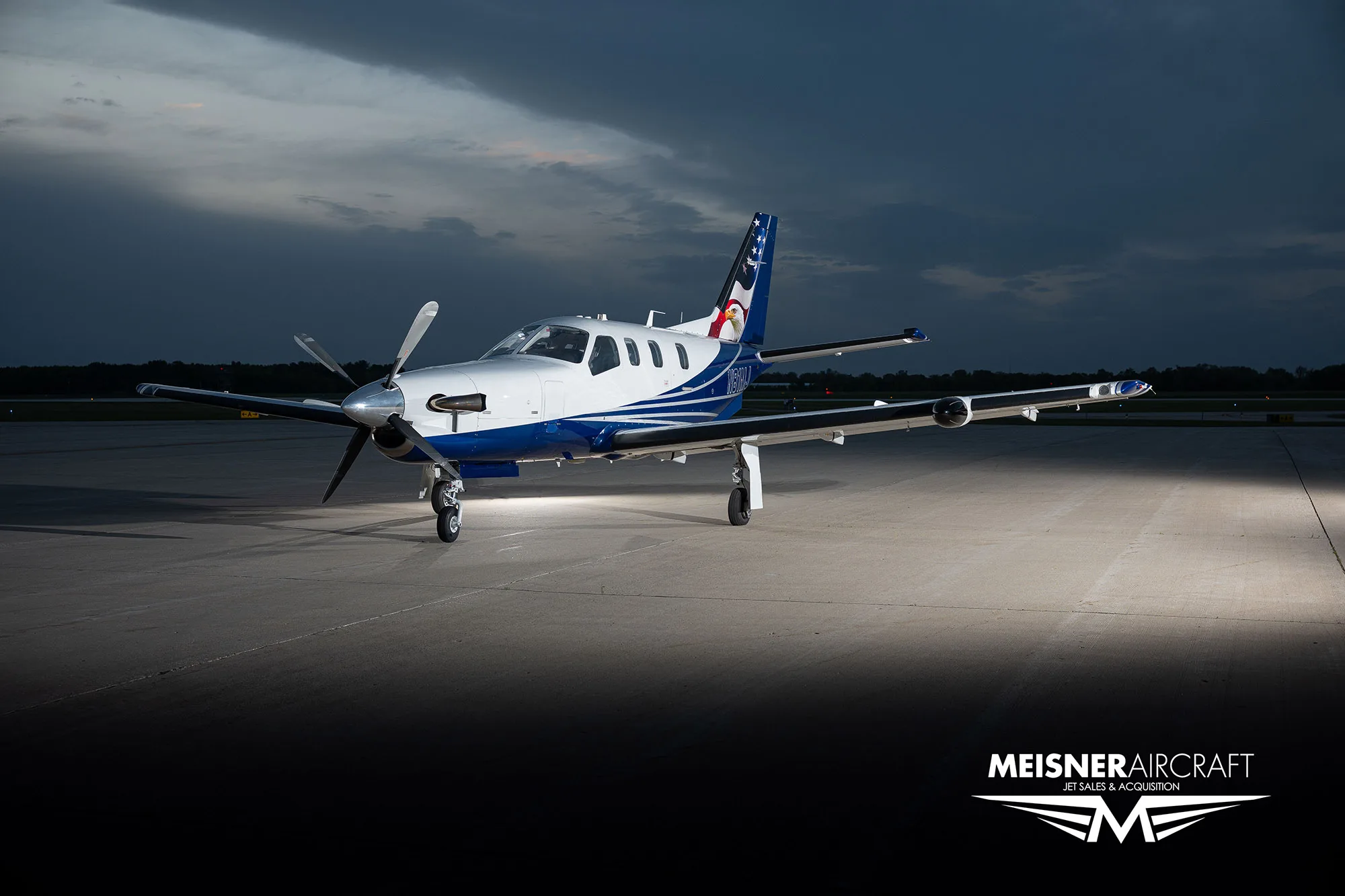 A TBM turboprop aircraft with a blue and white paint scheme and an eagle on the tail, parked on an airfield at dusk, viewed from the front-right.