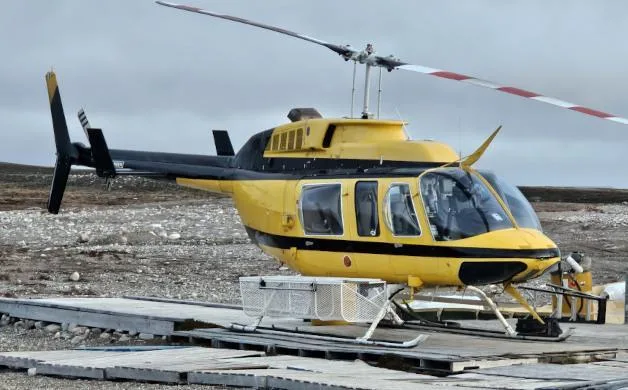 A yellow and black helicopter with a cargo basket on its skids, parked on a wooden landing pad under a cloudy sky.