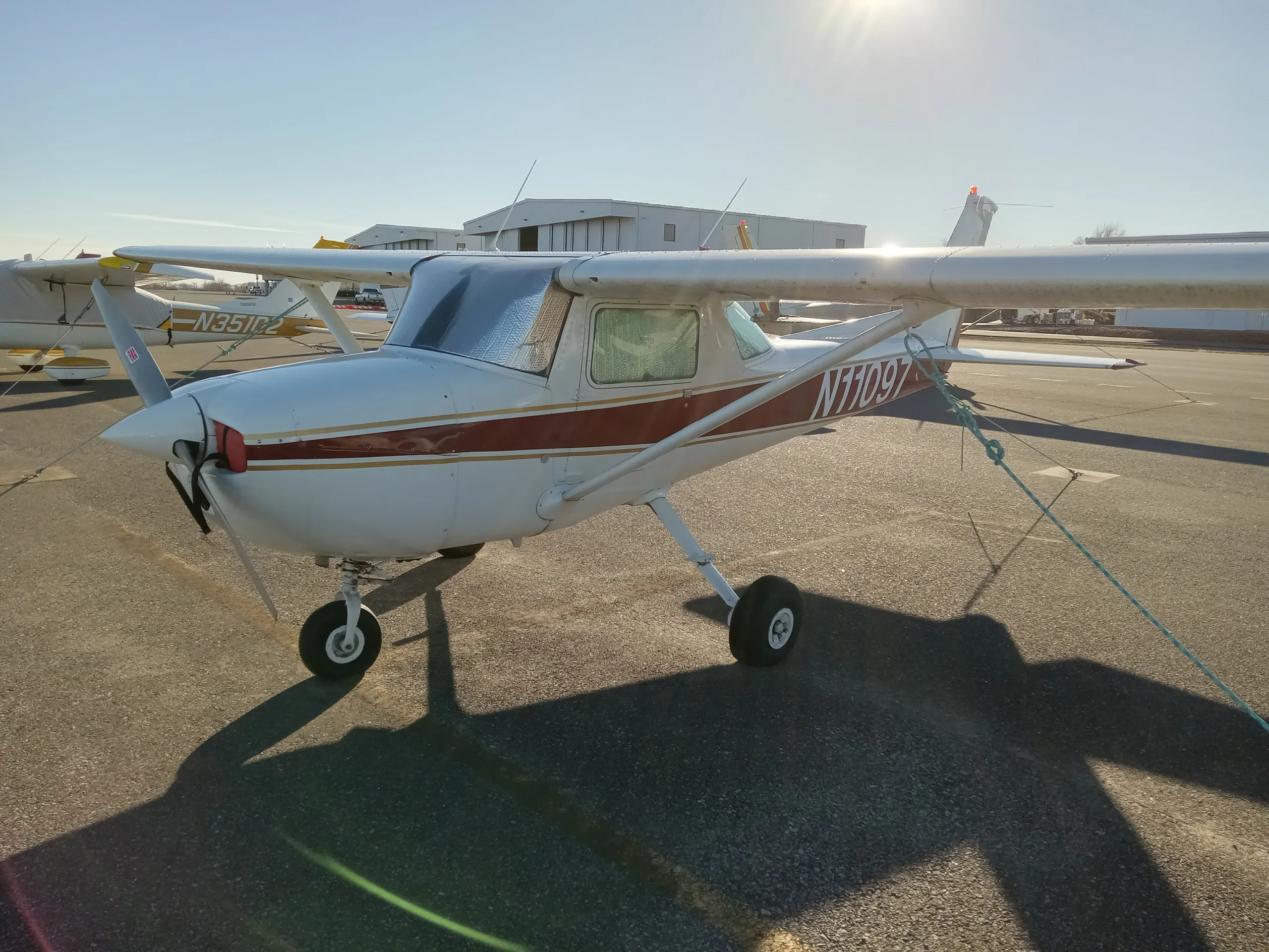 A white and maroon Cessna 150 or 152 aircraft, N11097, parked on an asphalt ramp with sun glare.