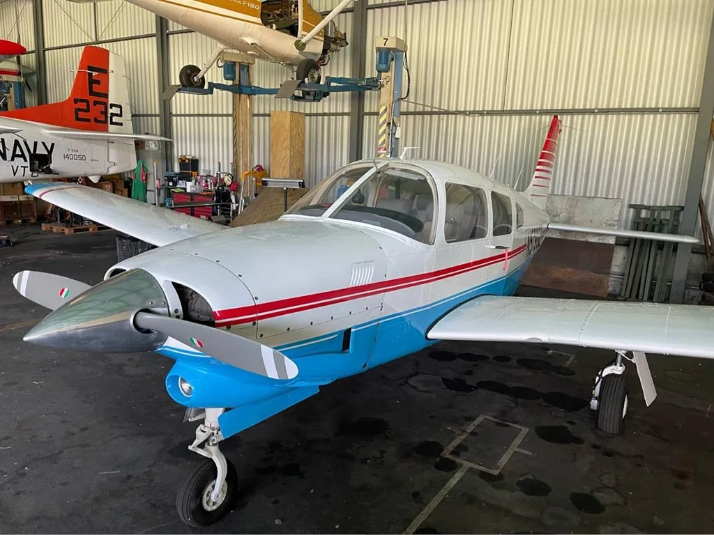 A front-left view of a white and blue Piper PA-28-181 Archer II with red stripes and Italian flag colors on the propeller, inside a hangar.