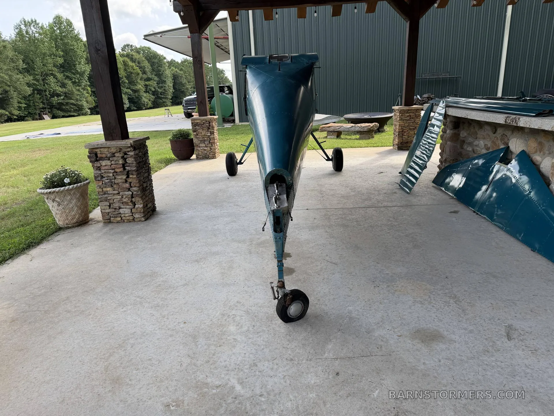 A rear view of the teal aircraft fuselage with its tail wheel, parked under a wooden shelter.