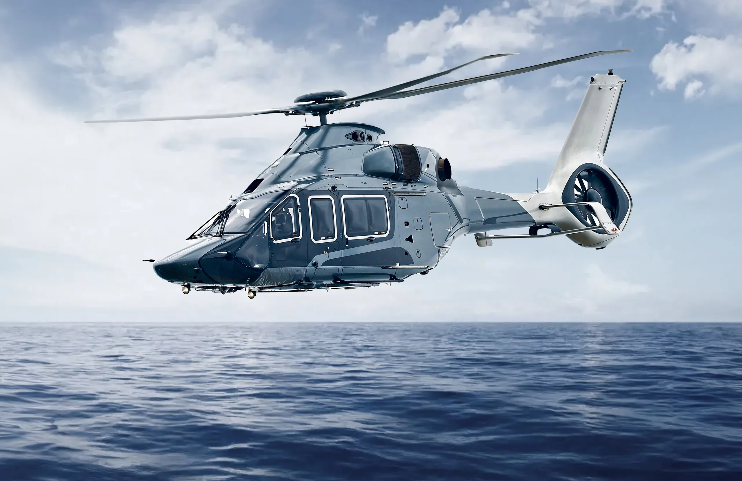A blue and white Airbus H160 helicopter flies over a calm blue ocean under a cloudy sky.