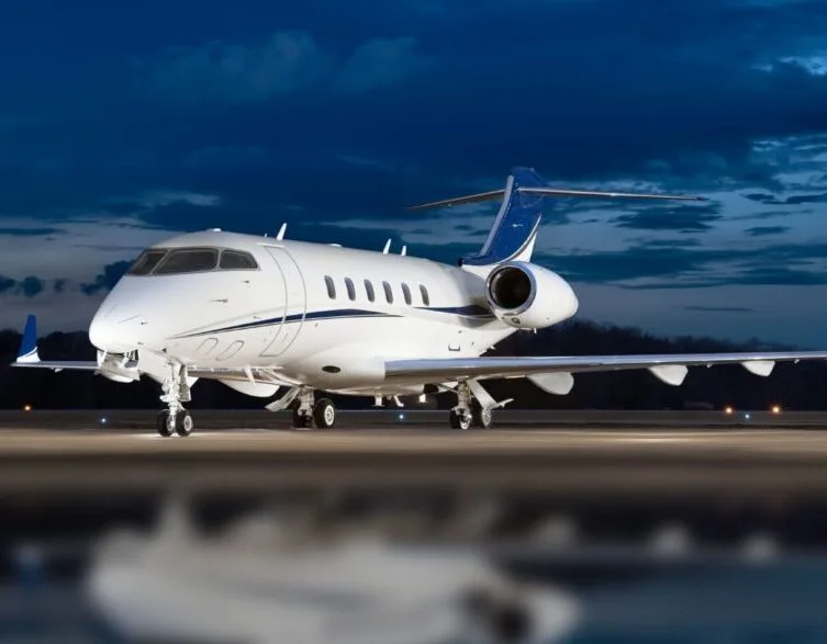 A white business jet with blue tail accents and stripes, parked on an airfield at dusk.