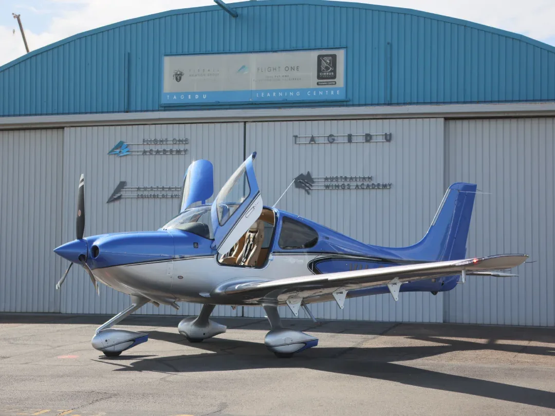 A blue and silver Cirrus SR22 aircraft with its gull-wing doors open, parked on an tarmac in front of a light blue and white hangar.