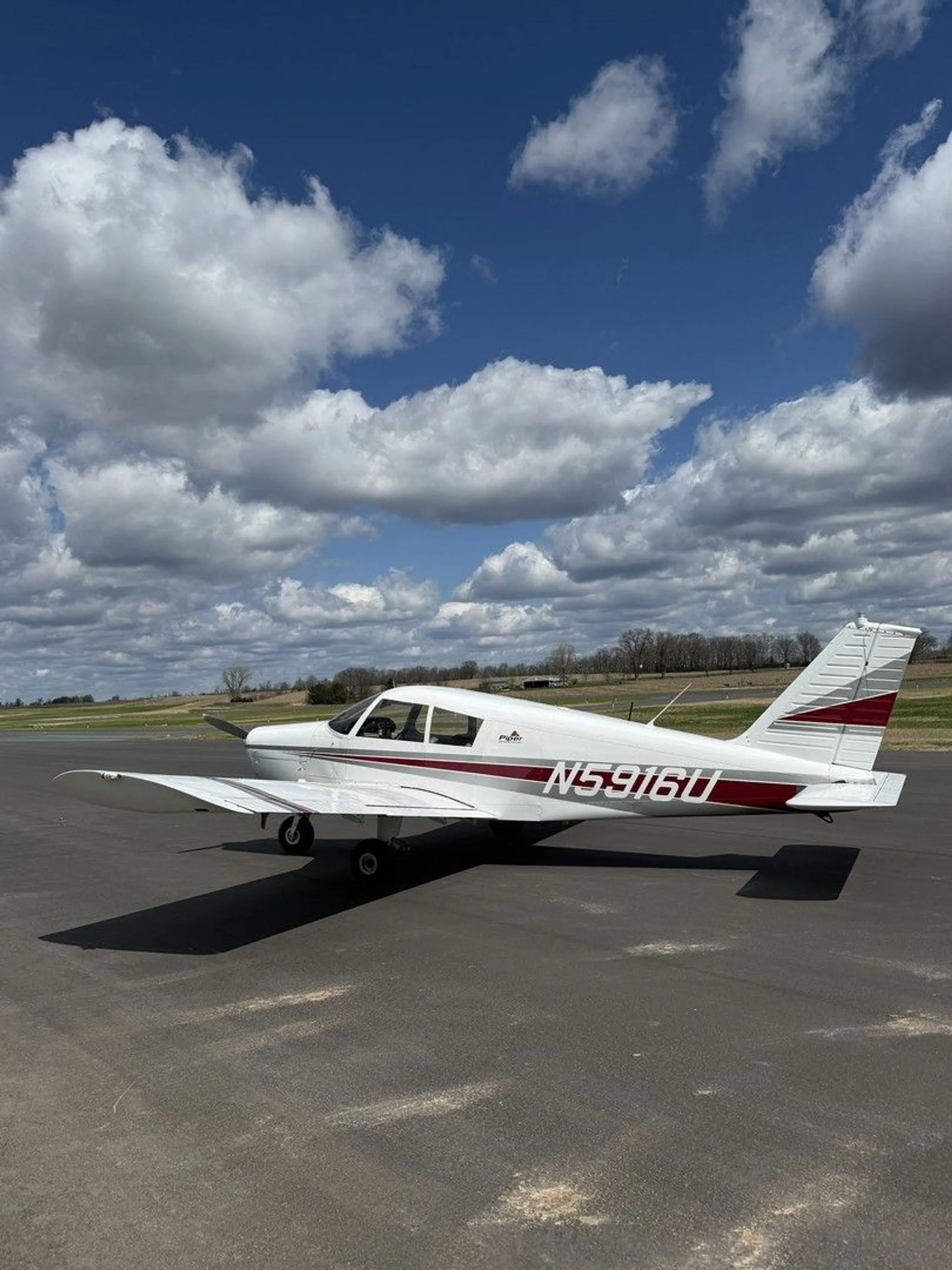 A white Piper Cherokee aircraft with red and grey stripes parked on an asphalt runway.