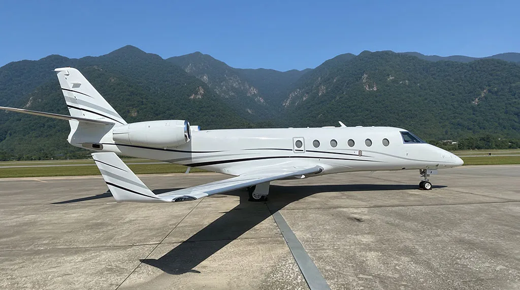 A white business jet with black and grey stripes is parked on an tarmac, viewed from the side.