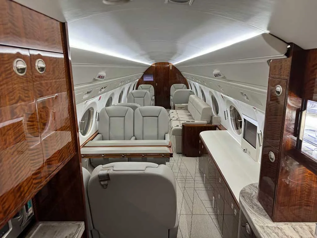 A wide-angle view of the aircraft cabin, featuring light gray seats, a wooden table, and glossy dark wood cabinetry.