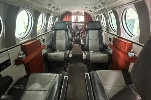 The aircraft's cabin interior features grey and black leather seats, wooden side tables, and round windows, looking towards the cockpit.