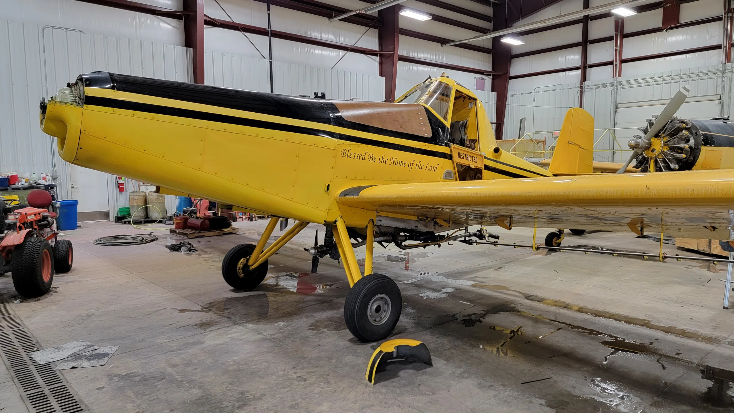 A yellow and black crop duster aircraft, N4941X, with 'Blessed be the Name of the Lord' and 'RESTRICTED' text, parked inside a hangar.
