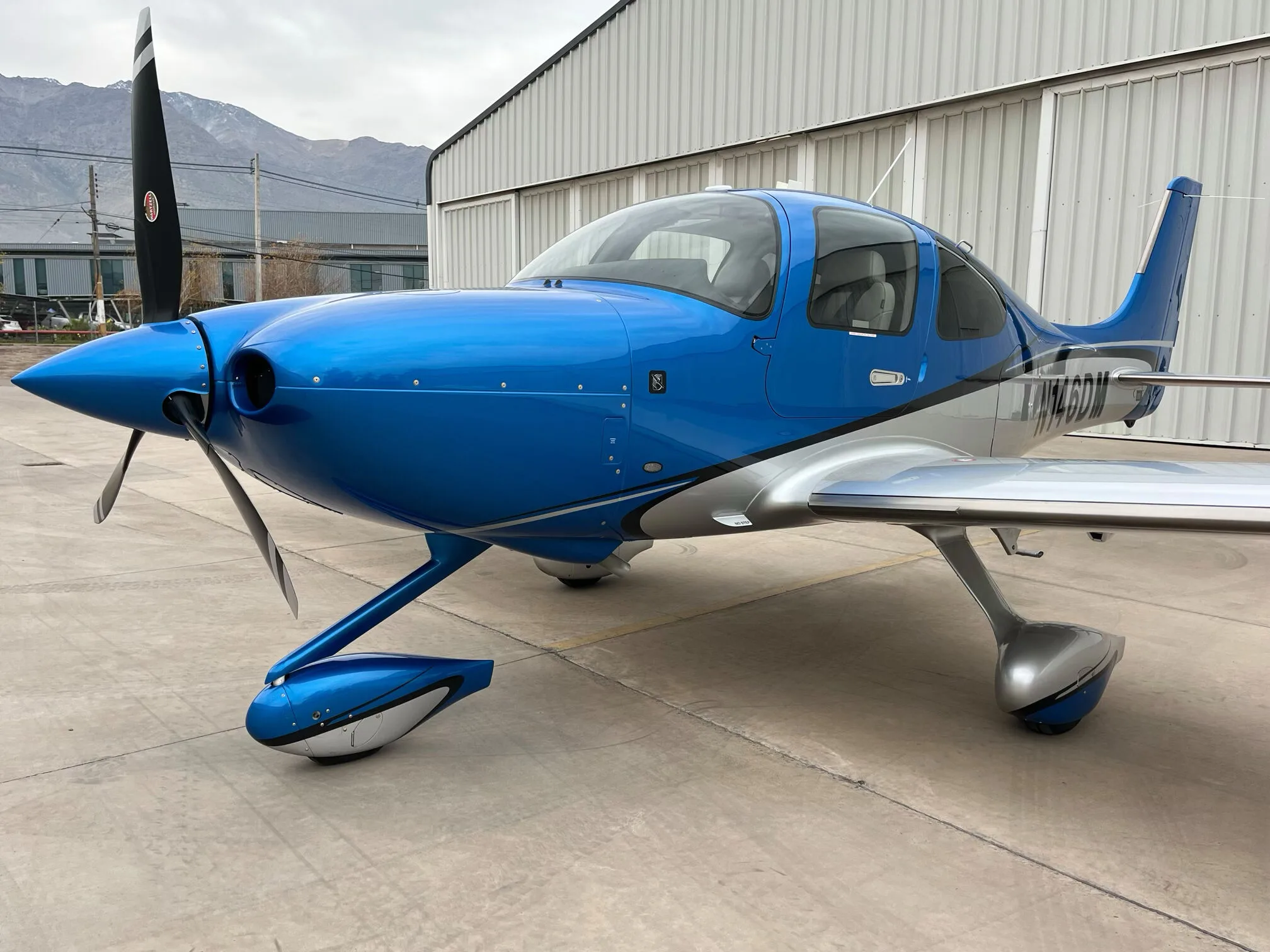 A front three-quarter view of the blue and silver Cirrus SR22 GTS aircraft, with mountains in the background.
