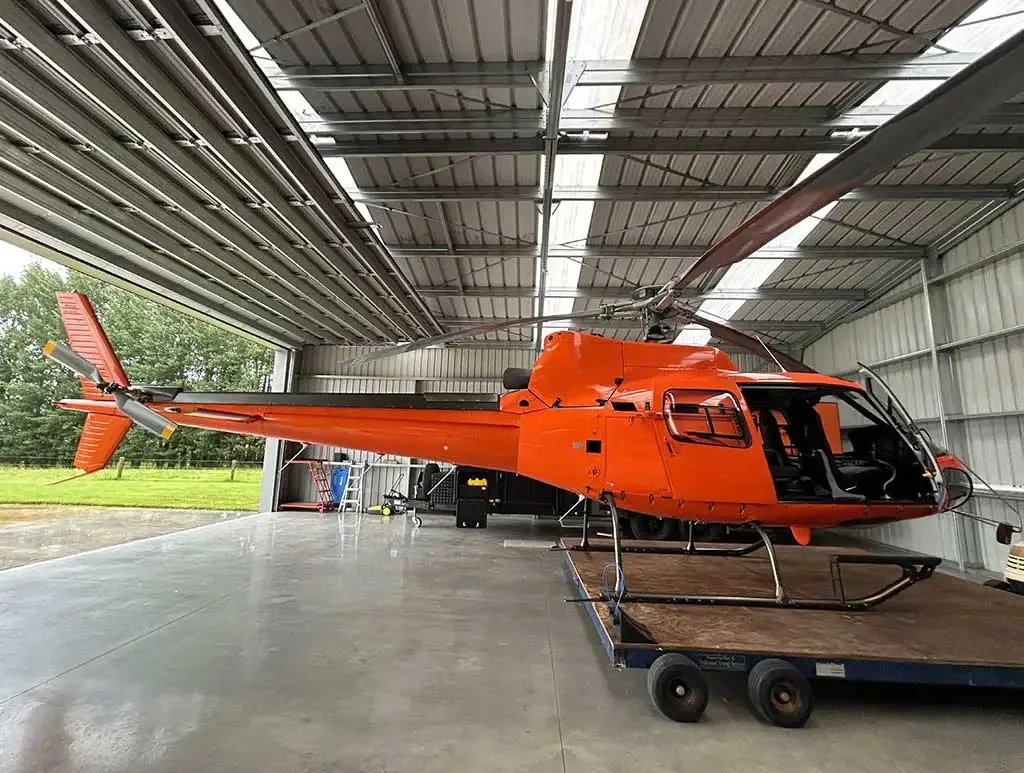 A bright orange helicopter with its main rotor blades extended, parked inside a metal hangar.