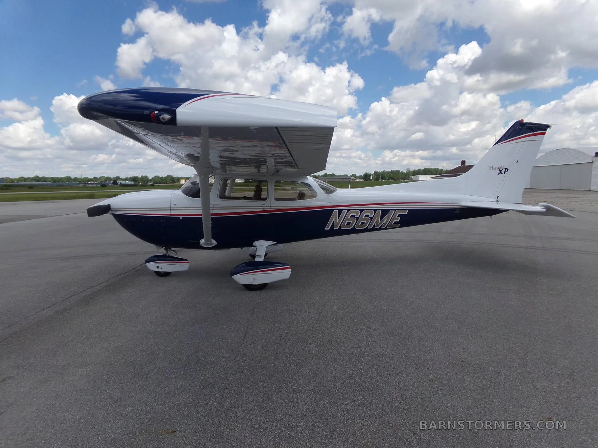 A white and blue Cessna 172 Hawk XP with red and gold stripes, seen from the side-right on an airfield under a partly cloudy sky.