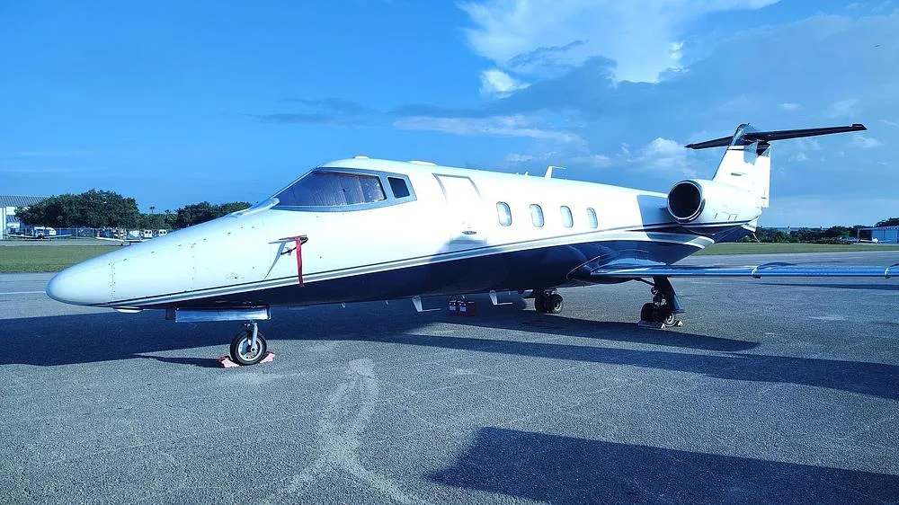 A white and dark blue private jet with its door closed, parked on an asphalt tarmac under a clear blue sky.