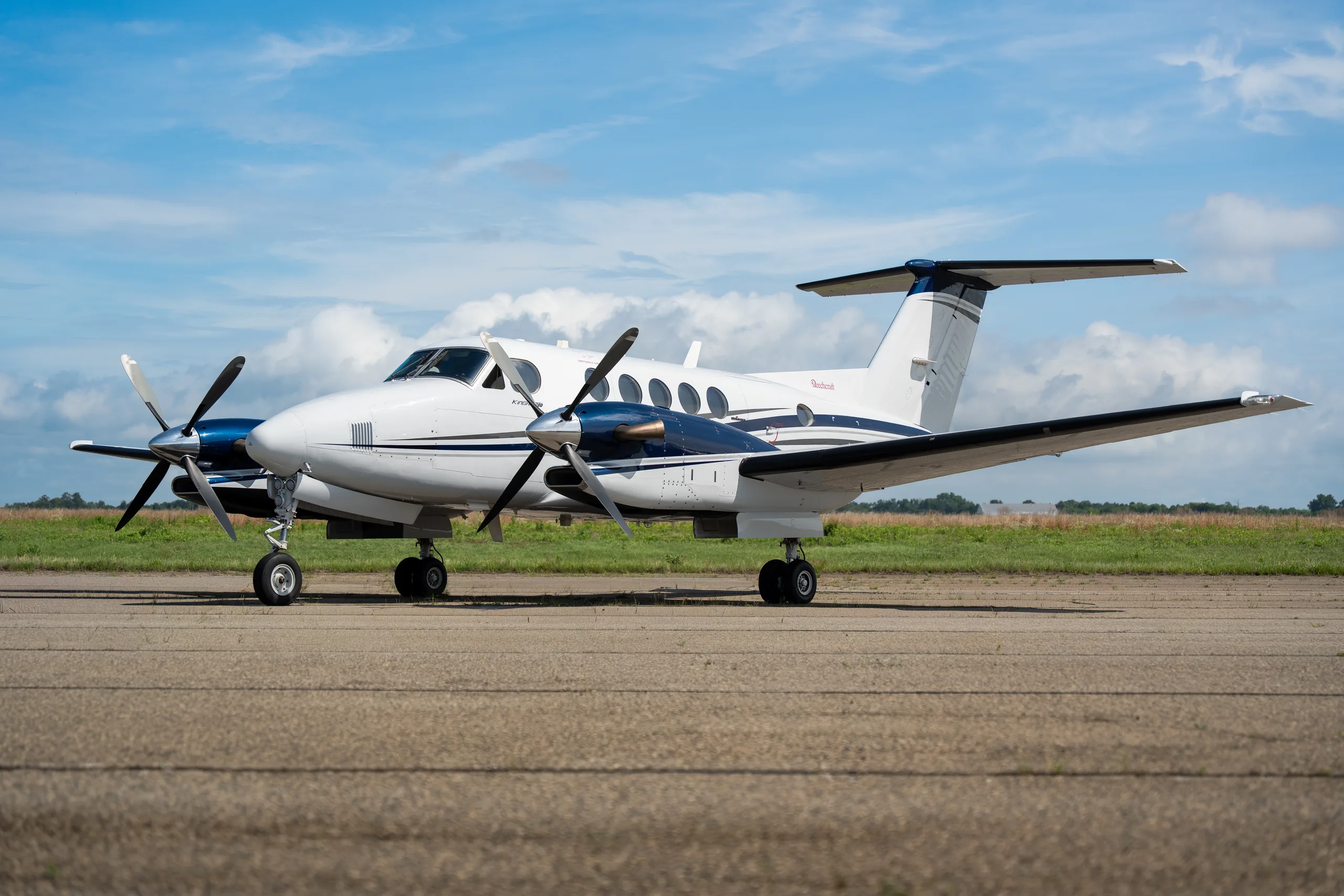 A white and blue Beechcraft King Air twin-turboprop aircraft parked on a tarmac under a partly cloudy sky.