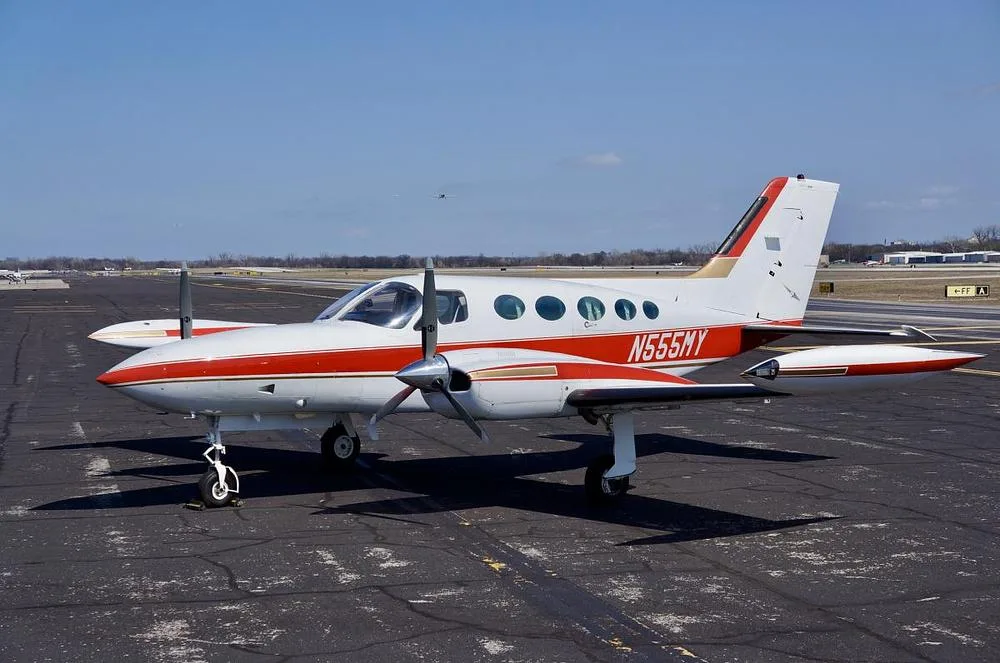 A white and red Cessna 400 series twin-engine aircraft, registration N555MY, is parked on an asphalt tarmac under a clear blue sky, viewed from the front-left.
