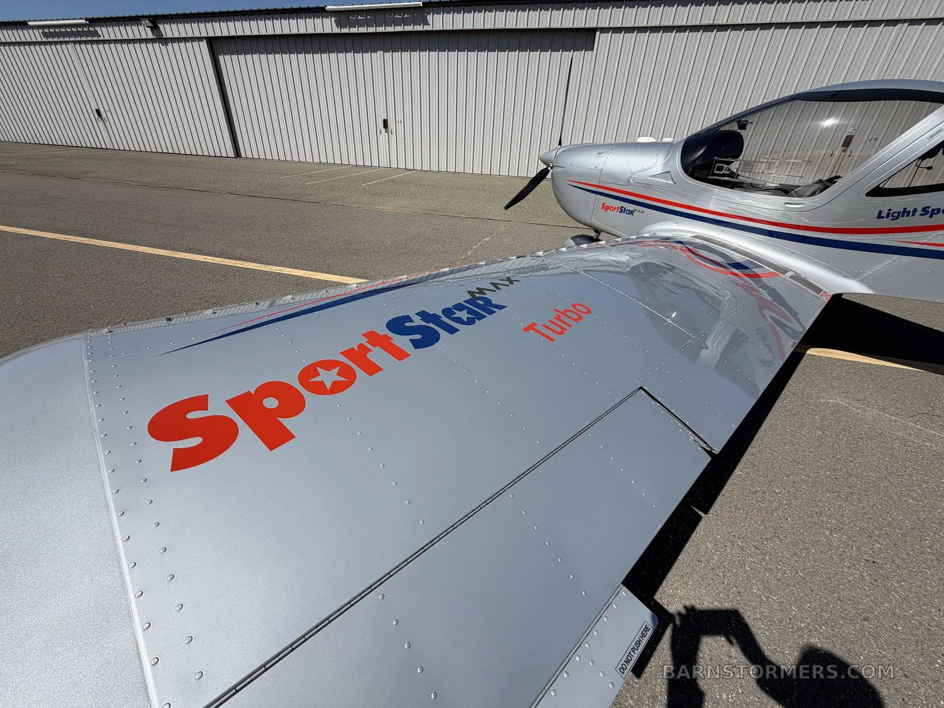 A close-up view of the silver SportStar MAX Turbo aircraft wing with red and blue stripes, parked on an asphalt tarmac.