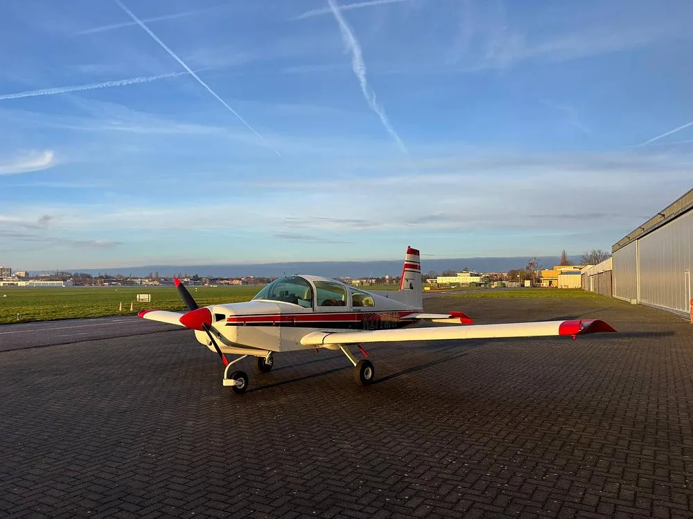A front-quarter view of a white Grumman American aircraft with red and black stripes, N277MW, parked on a tarmac near a hangar.