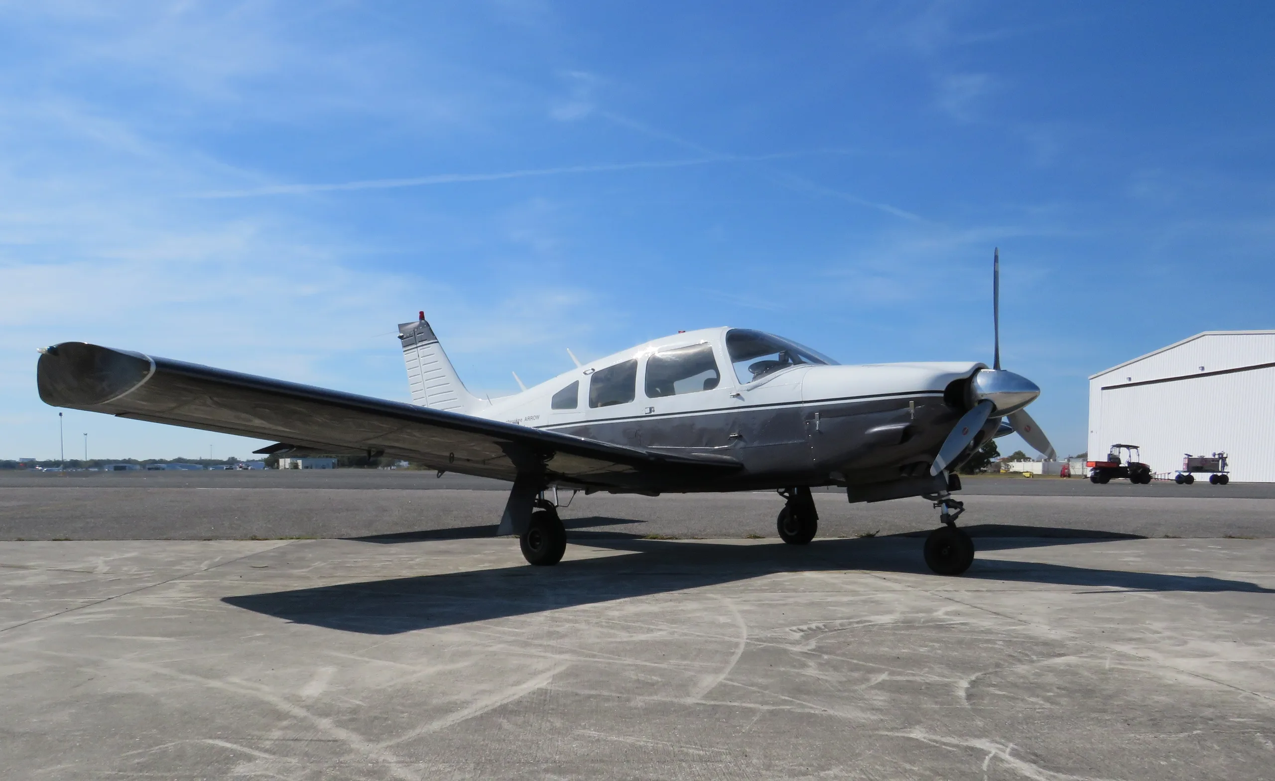 A white and grey Piper Cherokee Arrow single-engine aircraft is parked on an airfield under a clear blue sky.