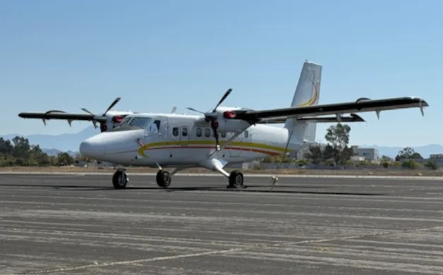 A white twin-engine turboprop aircraft with yellow and orange stripes is parked on an asphalt runway under a clear sky.
