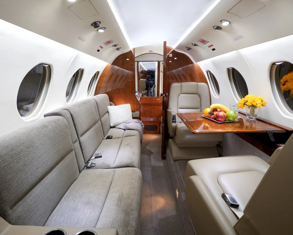 A wide view of the private jet cabin, showing a comfortable sofa, individual beige leather seats, and a wooden table with refreshments.