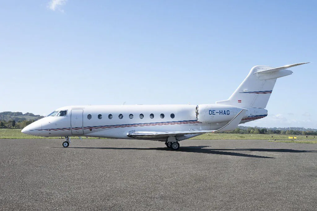 A white business jet with blue and red stripes, seen from the side, parked on an asphalt tarmac.