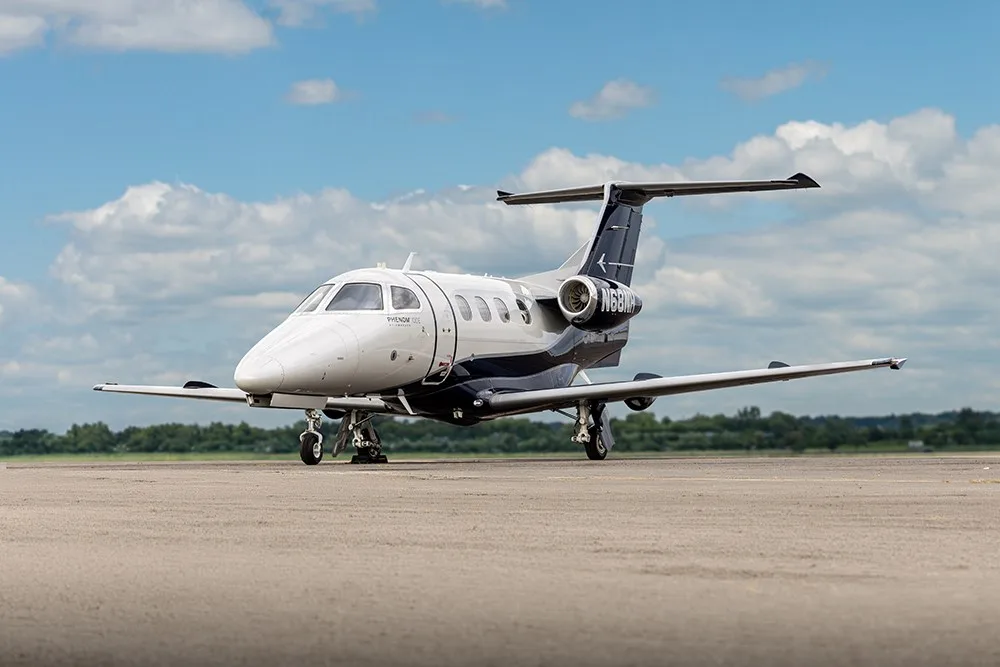 A white and dark blue Embraer Phenom 100E business jet, N68NH, is parked on an airfield under a partly cloudy sky.