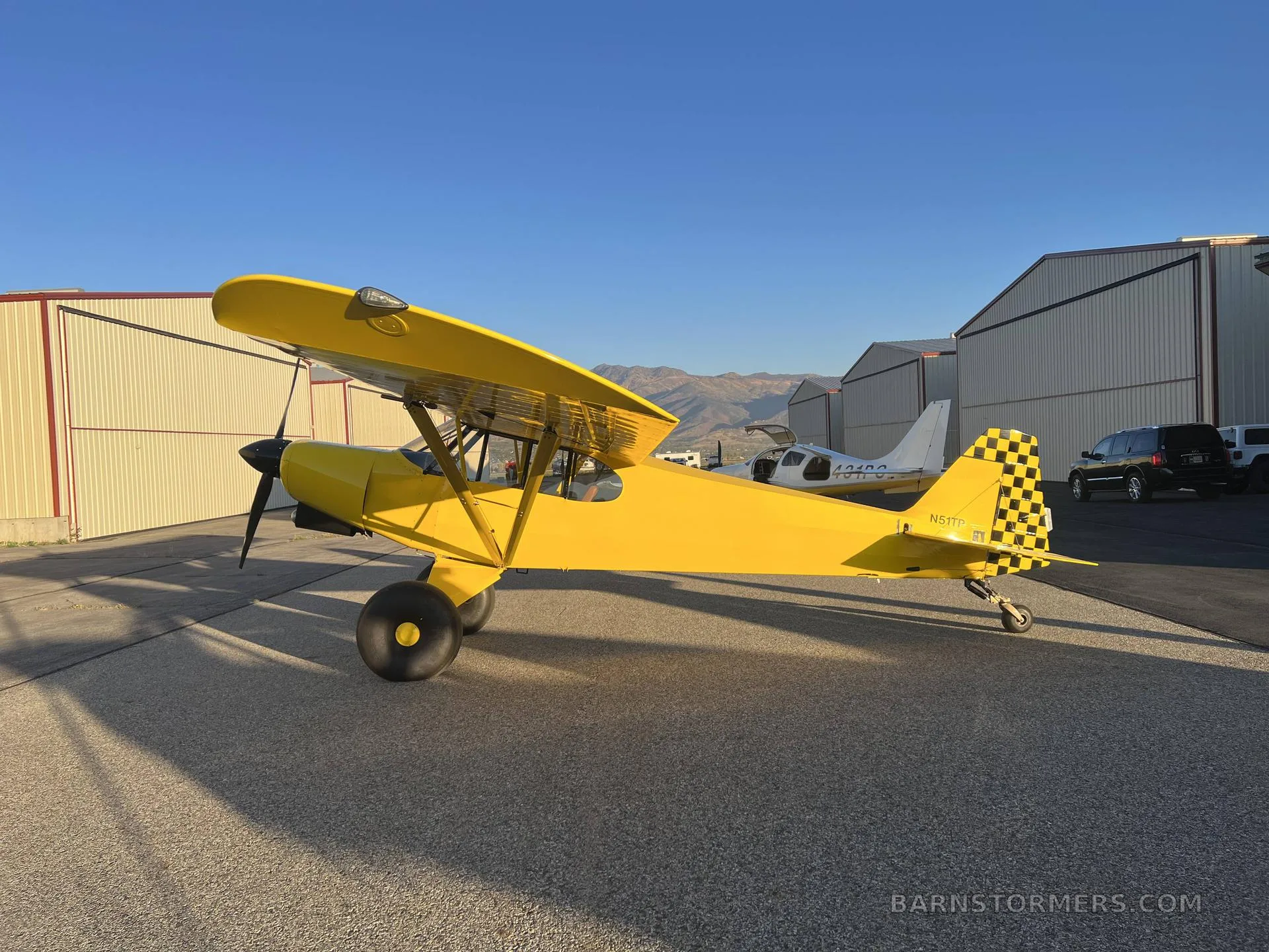 A bright yellow taildragger aircraft with a black and yellow checkered tail, parked on an asphalt ramp with hangars and mountains in the background.